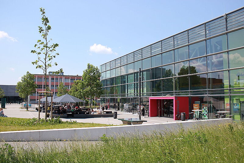 Gebäude mit Glasfassade, davor befinden sich Bänke und Tische mit Sonnenschirmen auf denen Menschen sitzen, sowie Gras und grüne Bäume