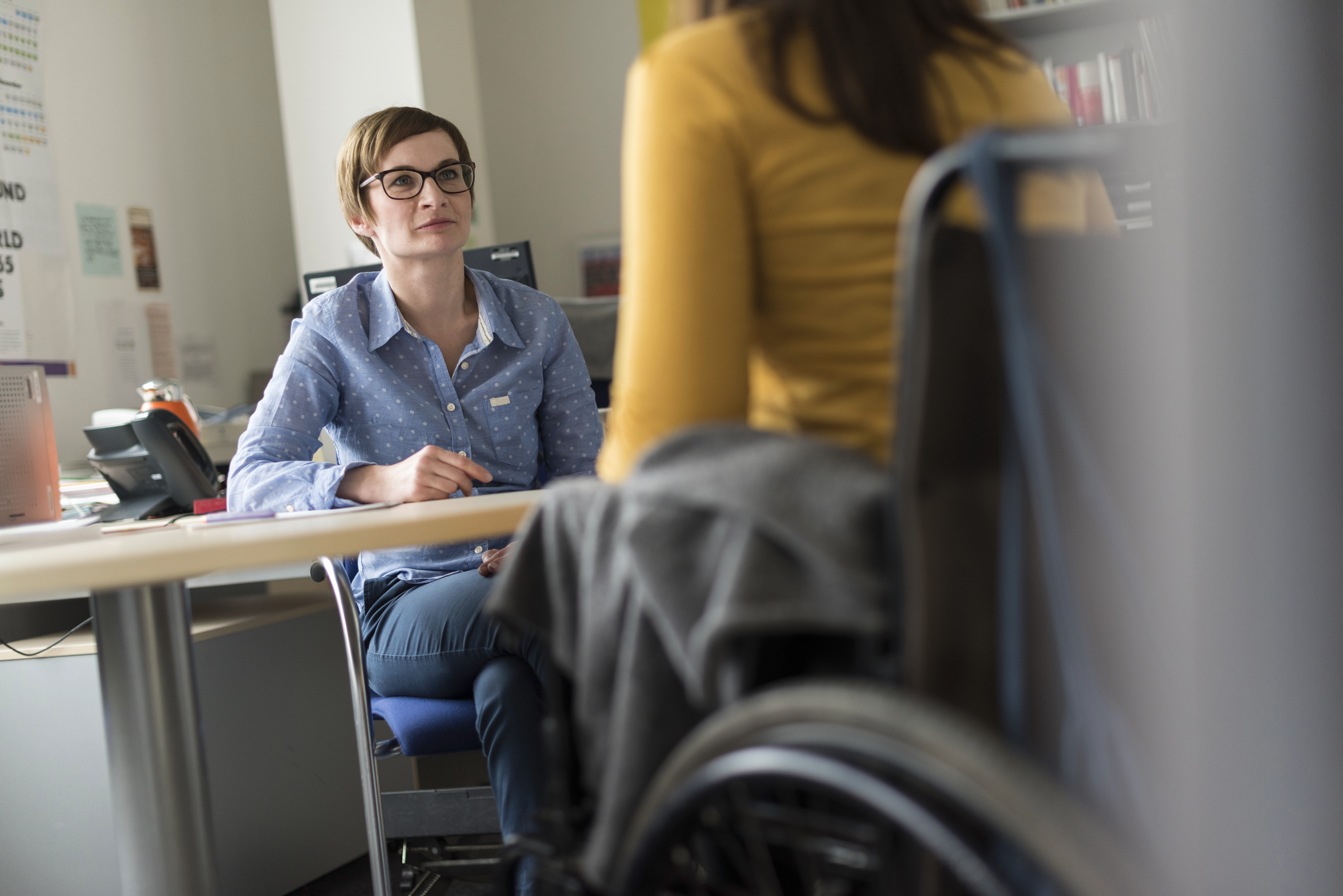 A woman (a counselor) and a woman in a wheelchair are sitting across from each other at a desk