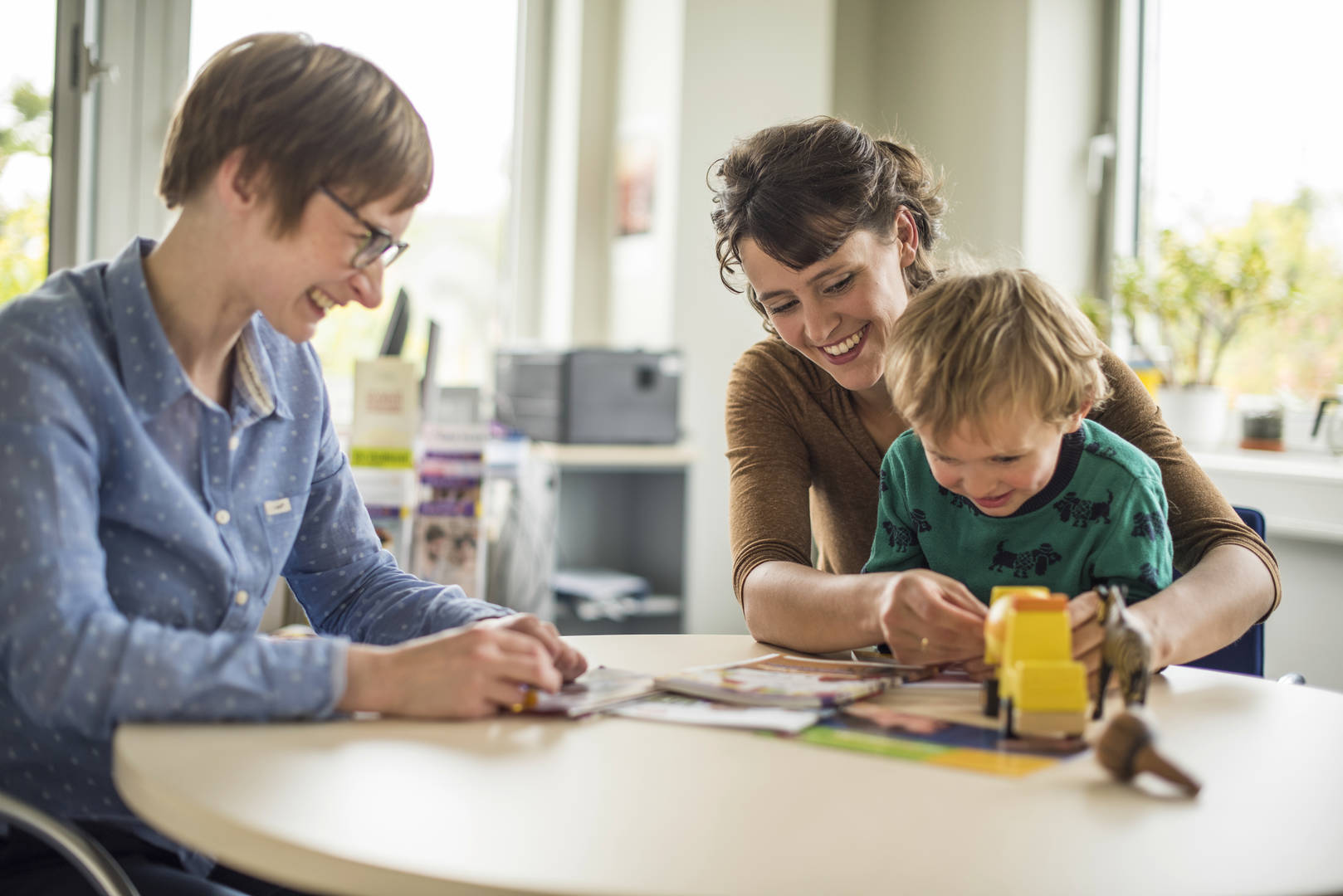 Zwei lächelnde Frauen und ein lächelndes Kind sitzen an einem Tisch und das Kind hat Spielzeug auf dem Tisch liegen und sitzt auf dem Schoß der einen Frau