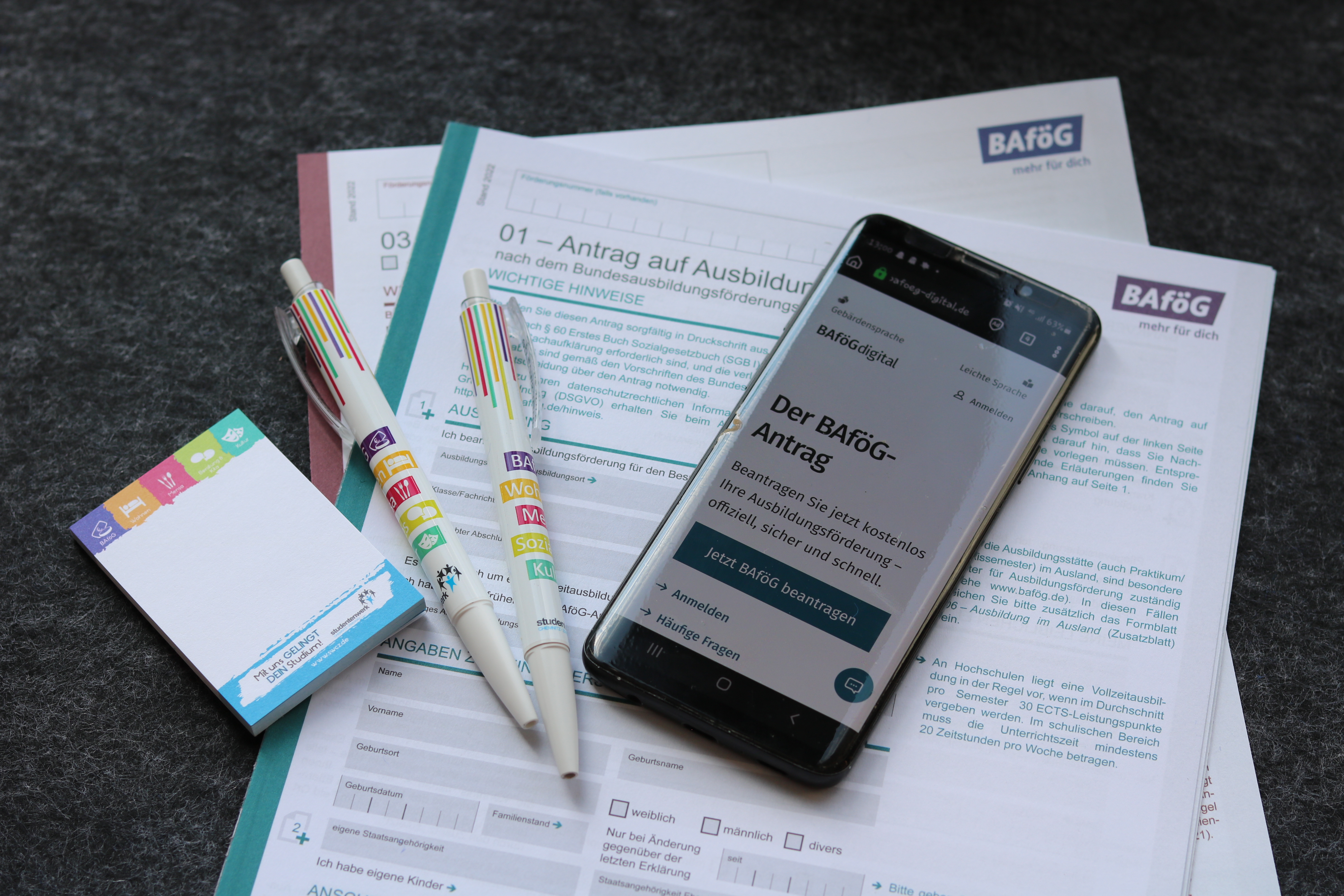 BAföG Anträge in Zettelform und ein Handy welches die Website bafög-digital zeigt