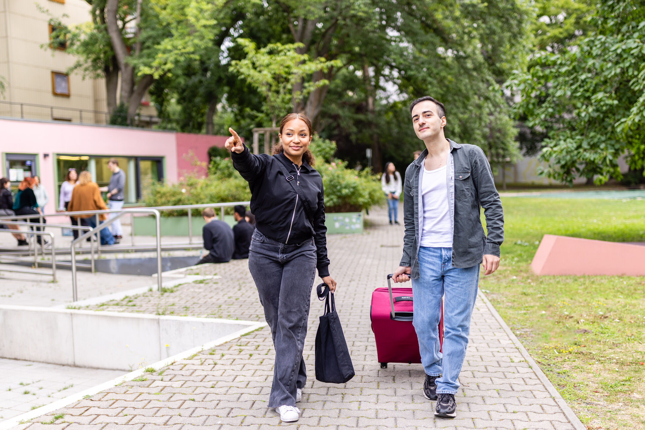 Ein Student mit Rollkoffer bekommt von einer Studentin die Richtung gezeigt