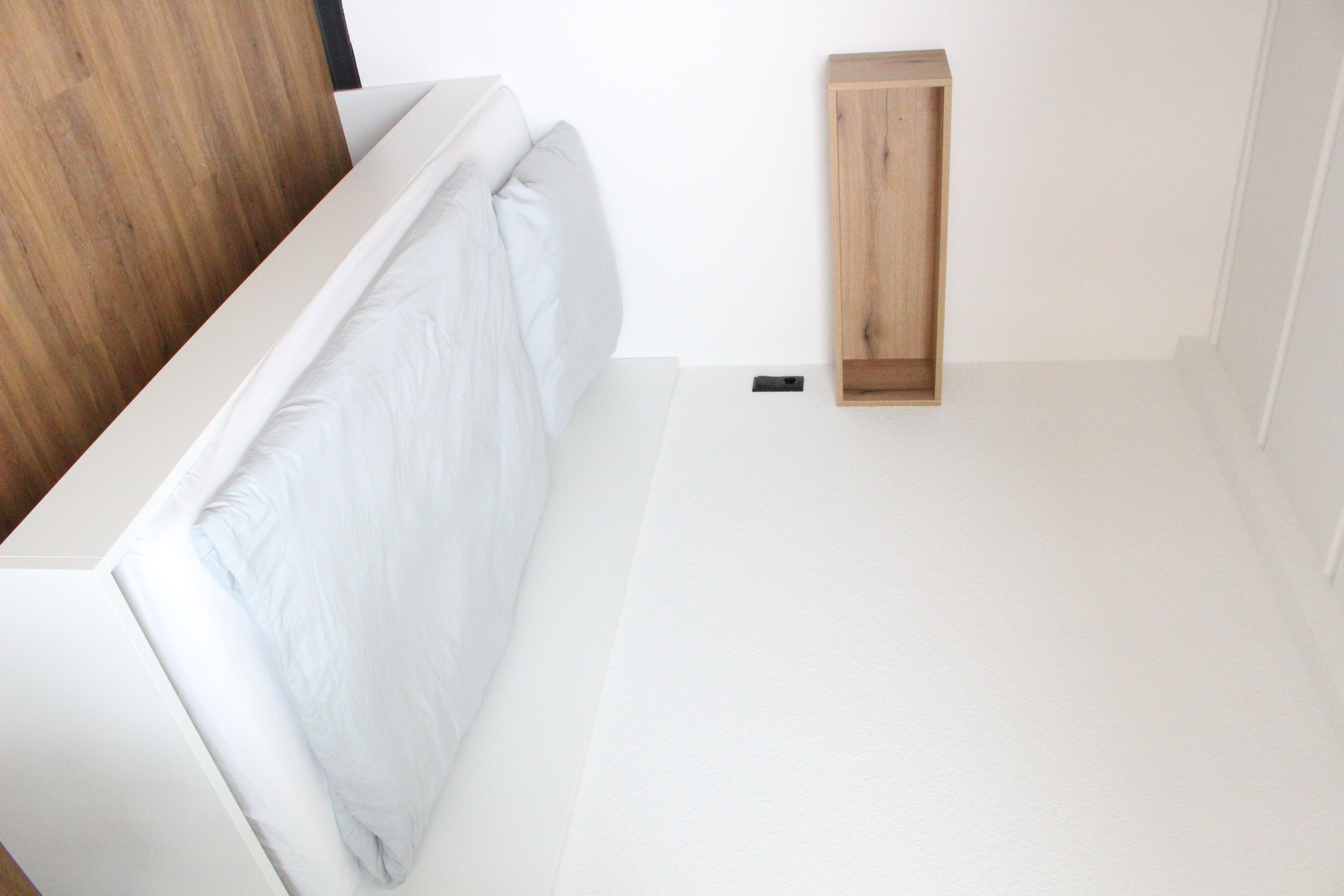 Weißes Bett mit heller Bettwäsche, dunkler Boden