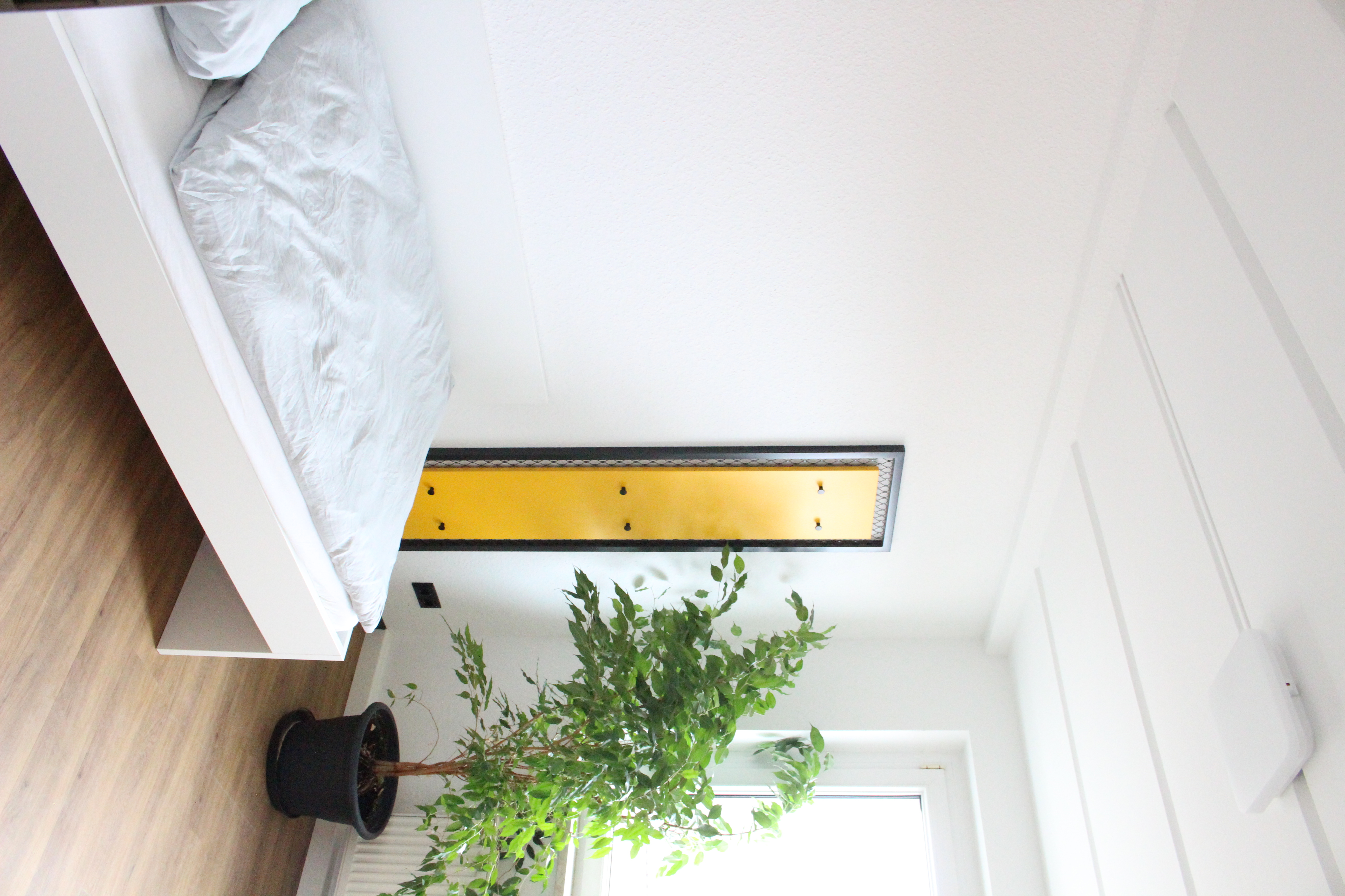 Weißes Bett mit heller Bettwäsche, gelbe Garderobe, große Pflanze